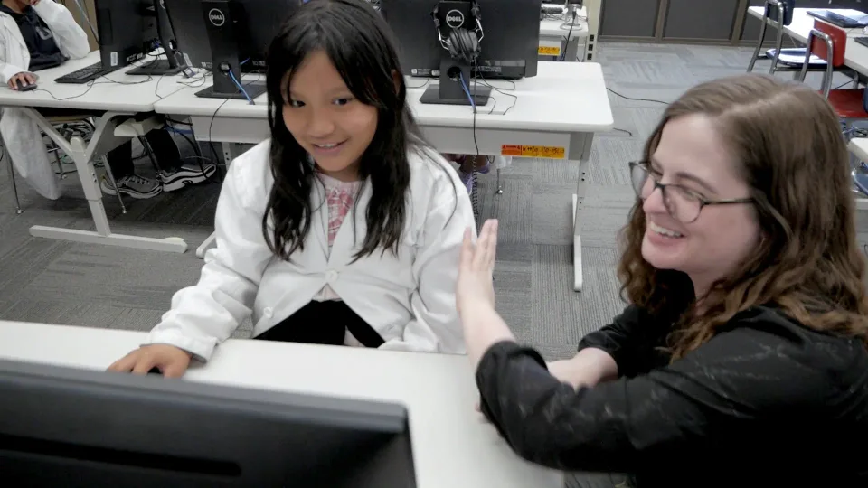 A teacher high-fives a student in a lab coat at her computer.