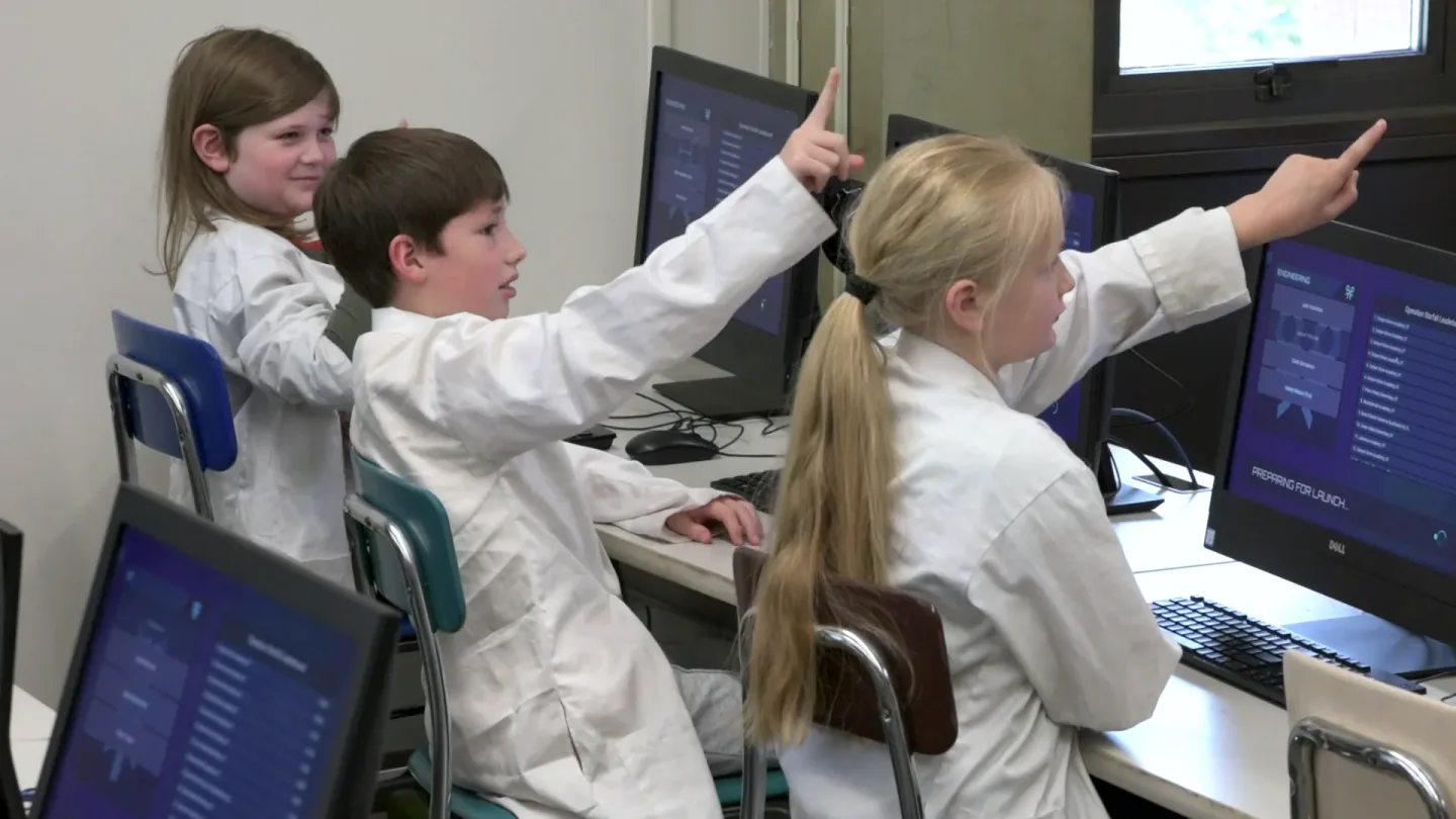 Three students in lab coats point up at the Mission screen, one talking as he points.