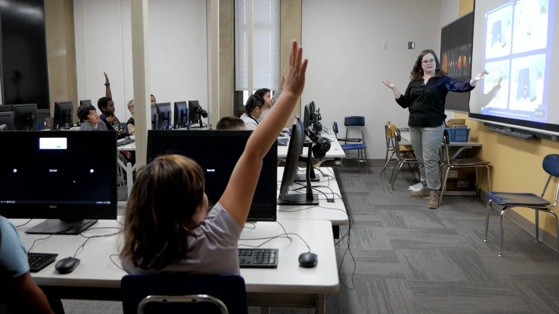 In a computer lab, a student's arm shoots up in the foreground while a teacher at the projected screen gestures to the class and more hands go up behind her.