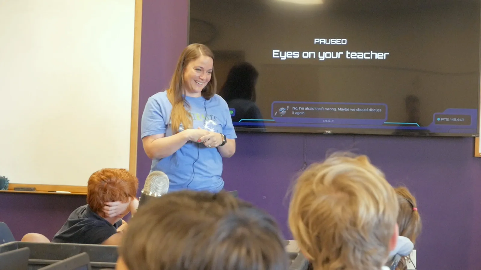 A teacher smiles at the front of the room while the Mission screen behind her reads Paused: Eyes on your teacher.