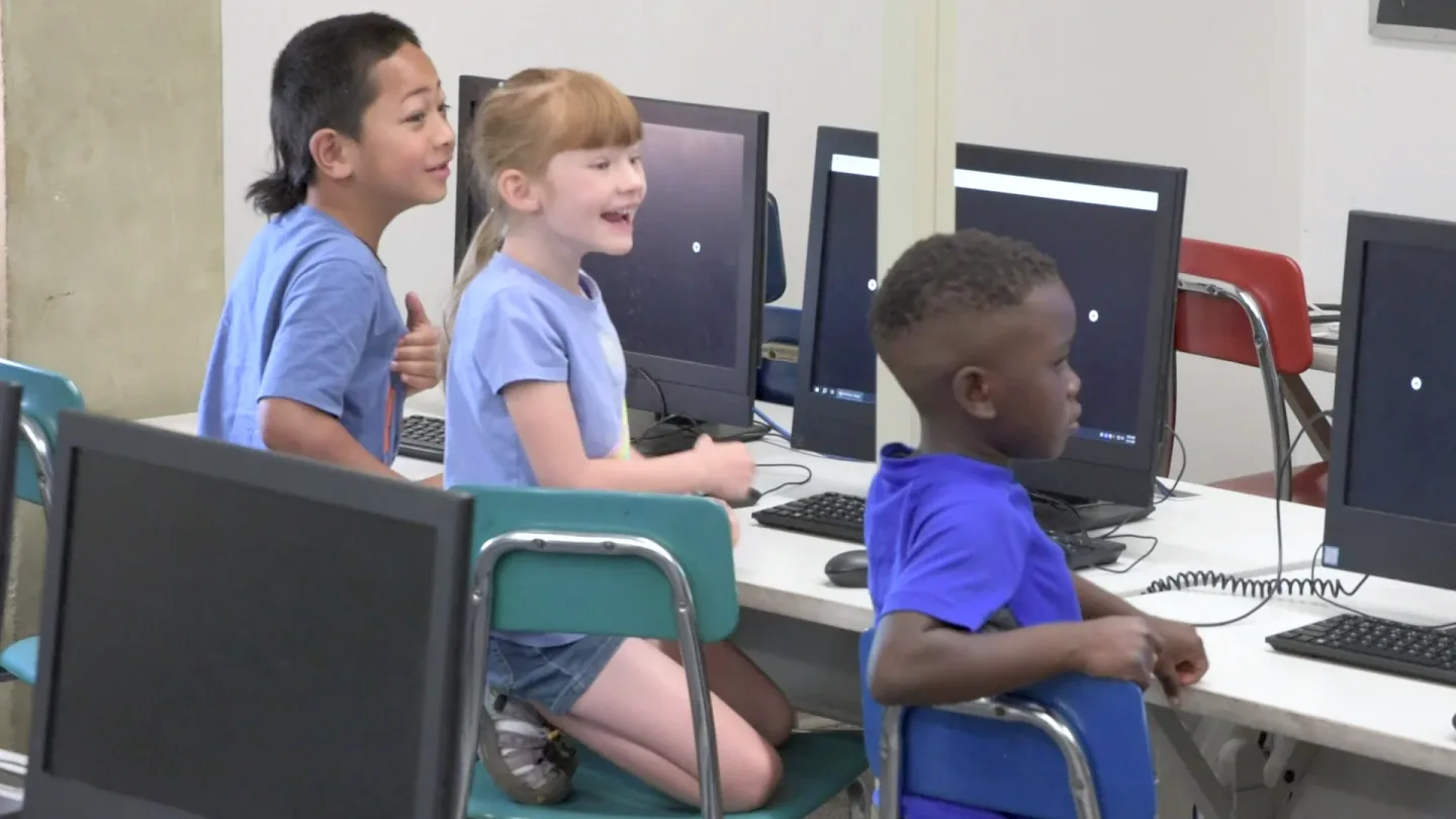 Three young students at lab computers look up at the wall screen, a girl laughing.