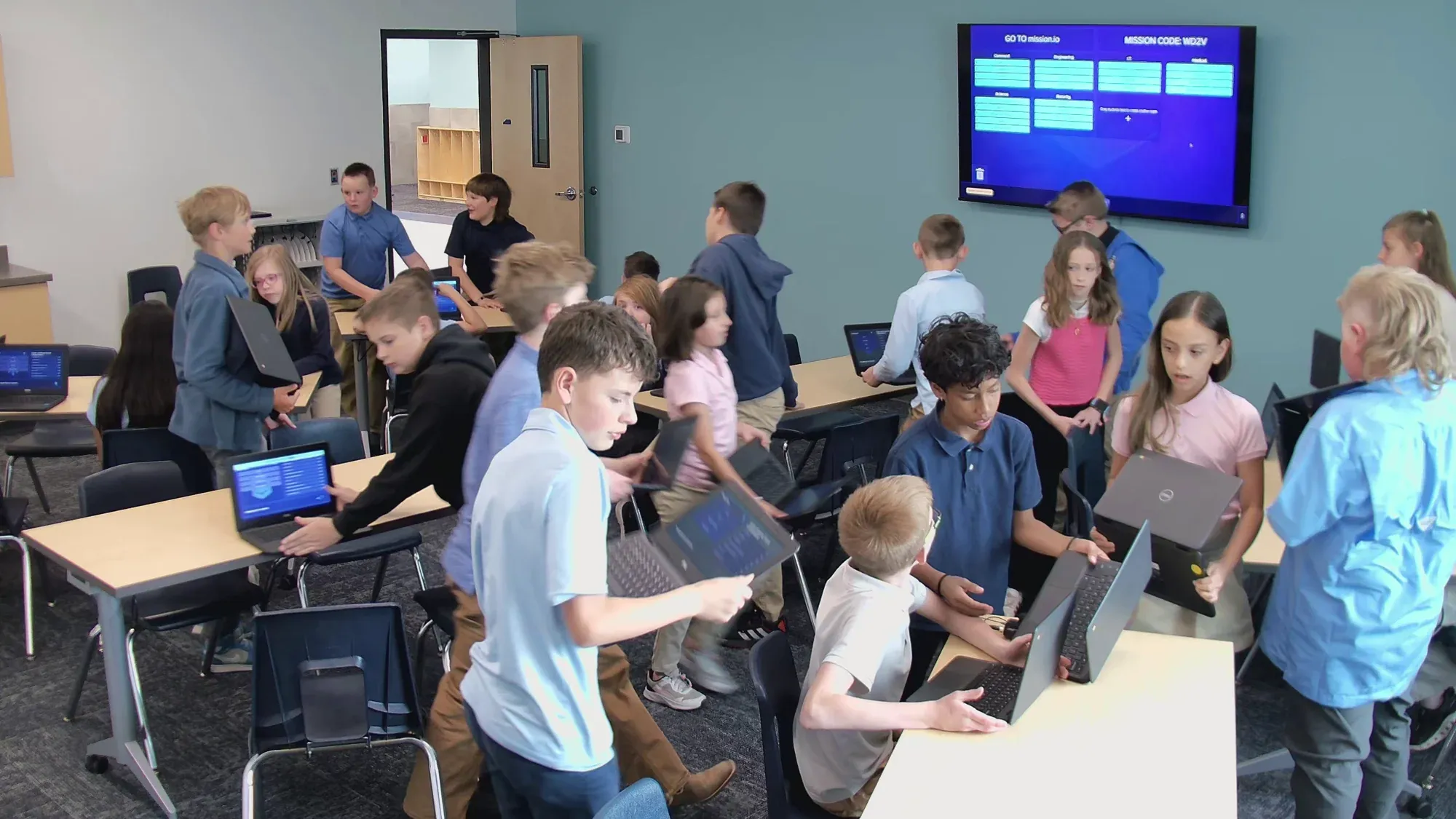 A class of upper-elementary students moves into crews with their Chromebooks while the screen at the front lists the stations.