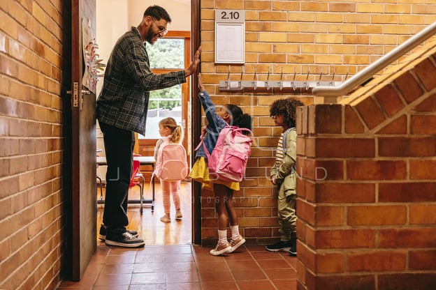 A smiling teacher at his classroom door high-fives a girl with a pink backpack as other students wait.