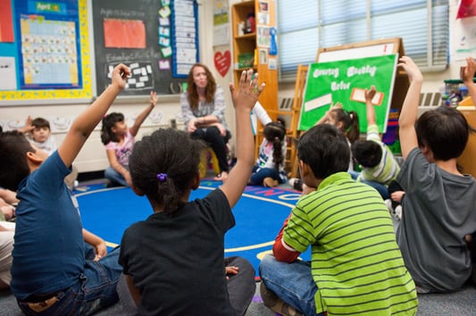 Young students sit in a circle on a rug in a classroom, many raising their hands, while a teacher sits across from them.