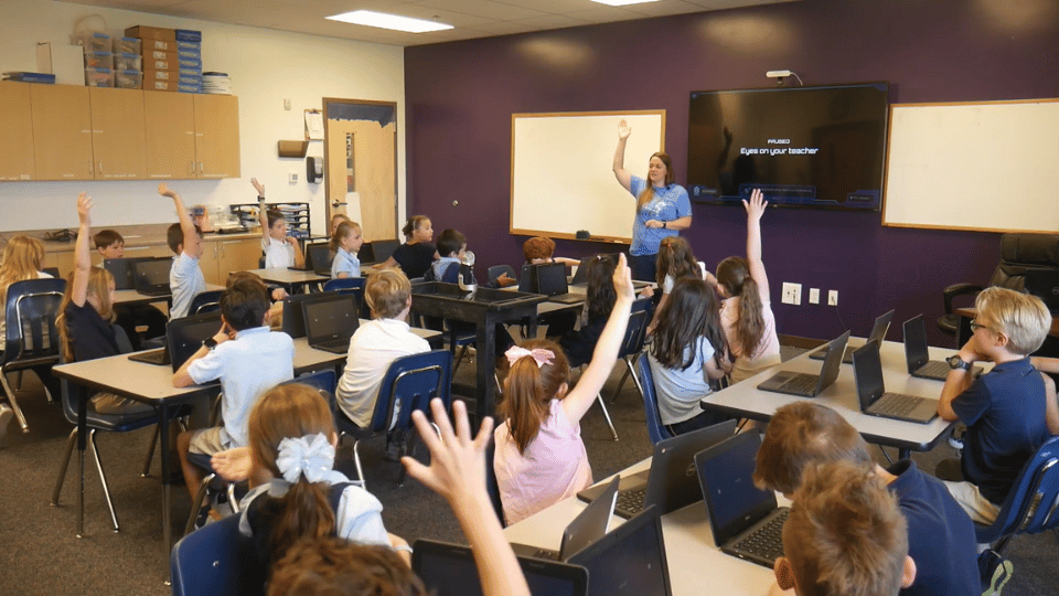 Elementary classroom of students raising hands while using laptops, engaging in critical thinking and interactive problem-solving with teacher guidance.