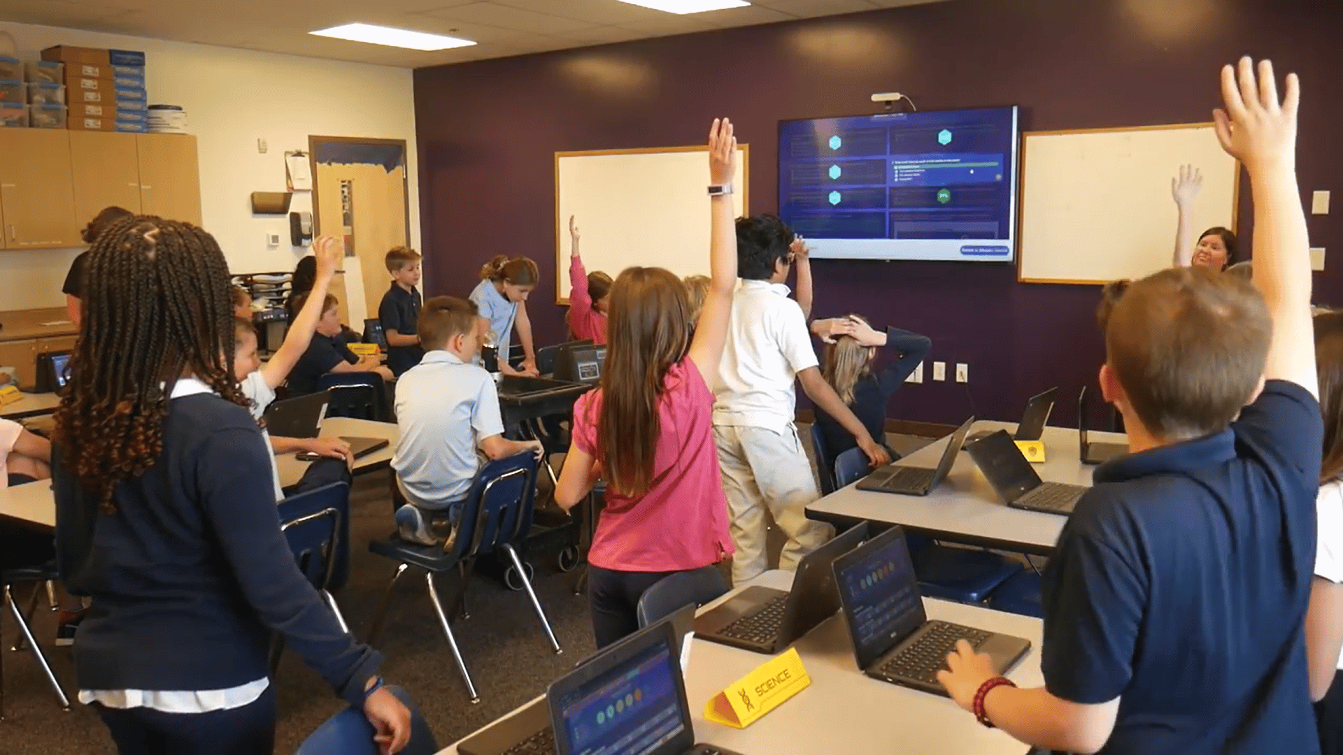 Students raising hands and engaging in critical thinking during a collaborative classroom mission, using laptops and interactive problem-solving to explore science concepts.