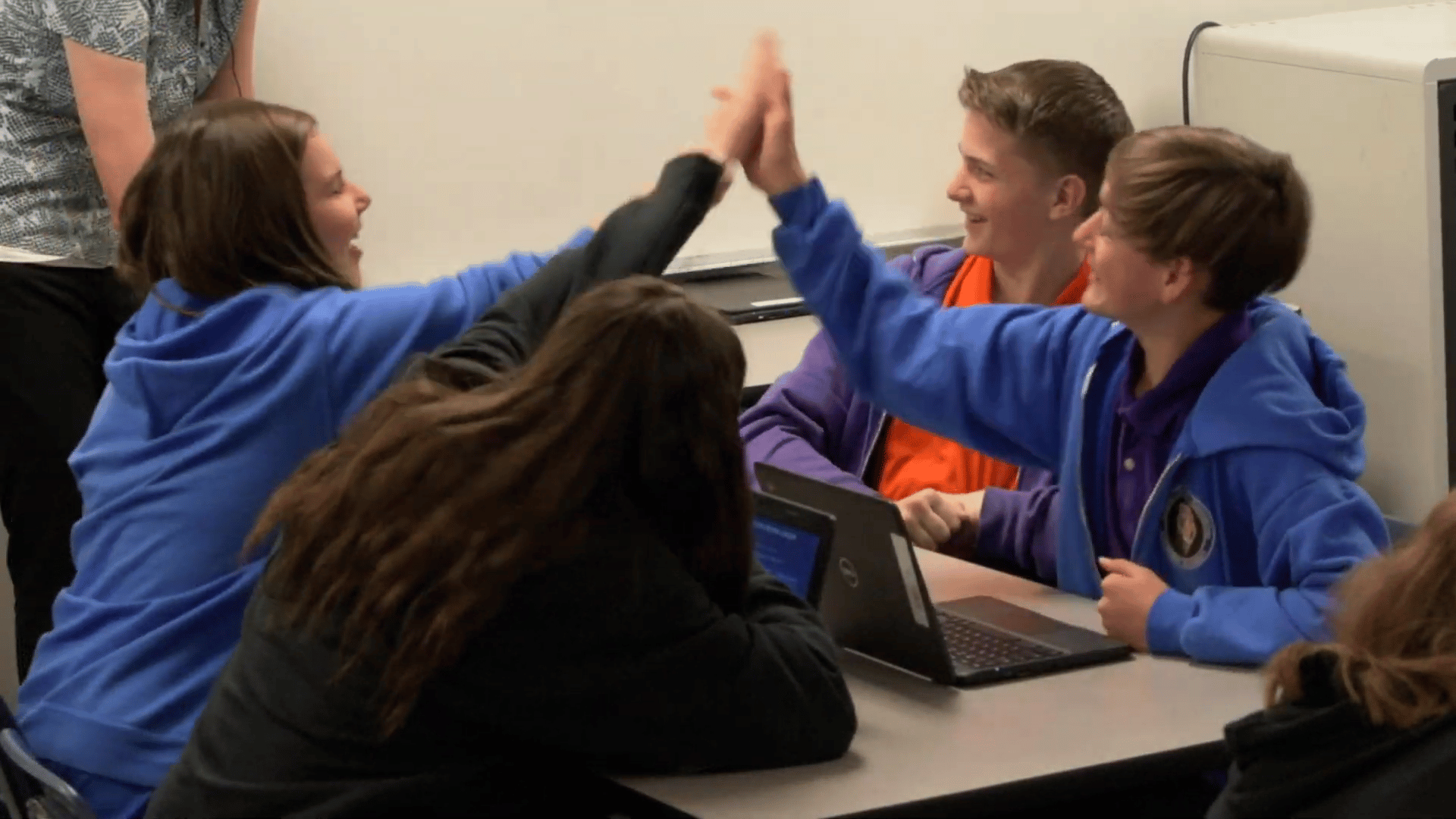 Middle school students celebrating with a high five after solving a problem together on laptops, showing collaboration, critical thinking, and teamwork in the classroom.
