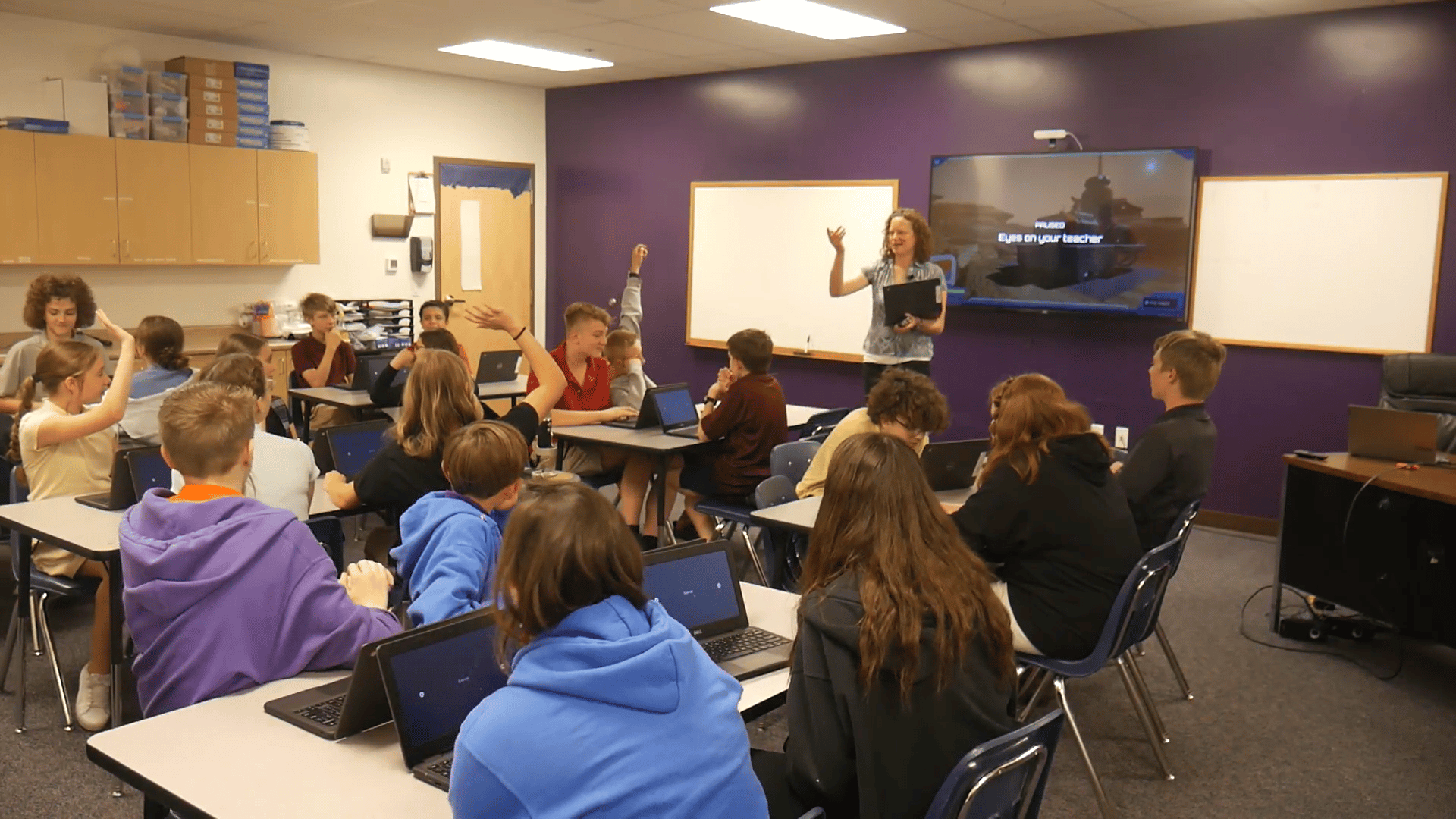 Teacher guiding a classroom mission as students raise their hands and discuss solutions, using laptops and critical thinking to solve interactive challenges together.